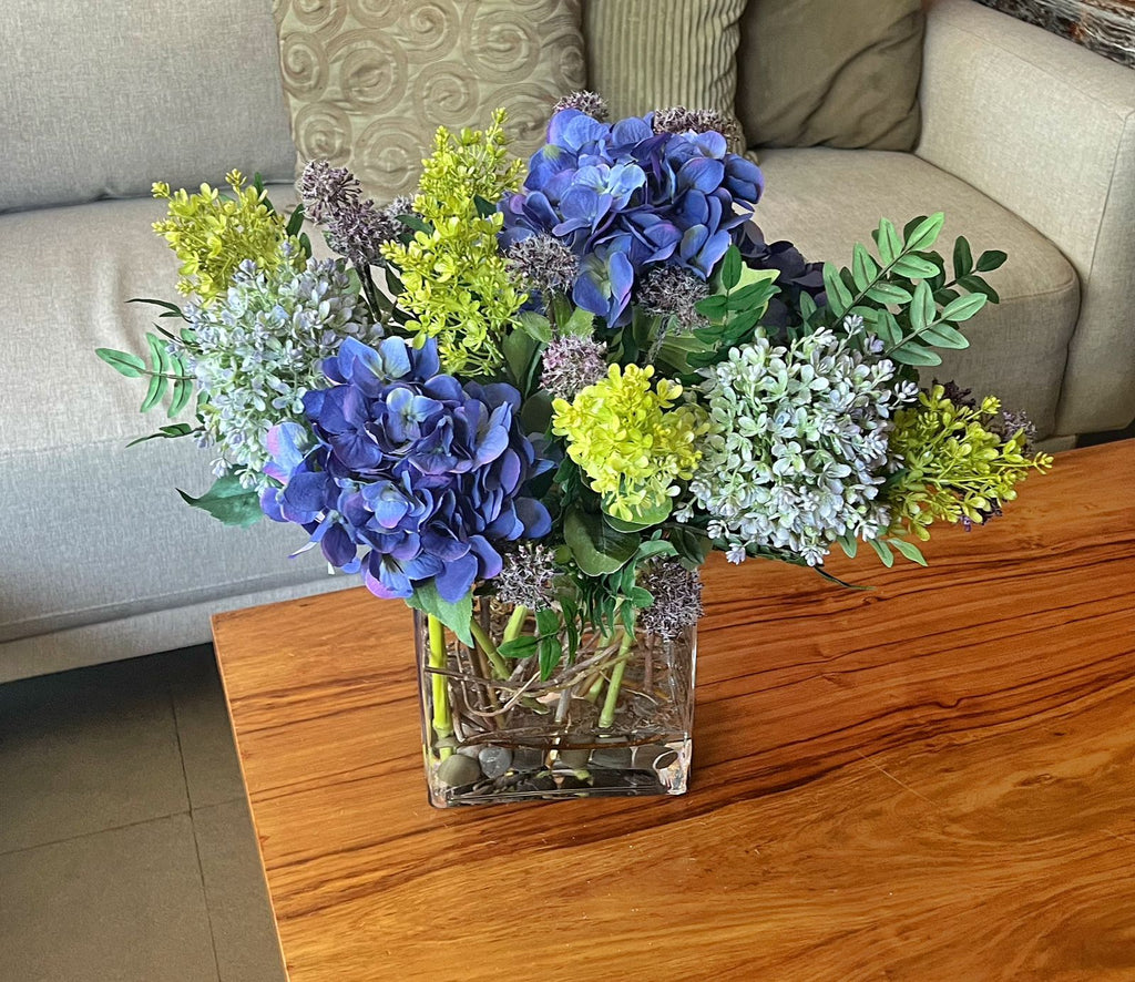 Arreglo Floral de Hydrangeas en Tonos Lila y Follajes Artificiales en Base de Vidrio con Agua Acrílica