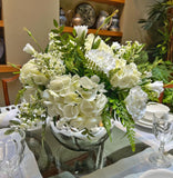 Centro de Mesa con Hydrangeas en Tonos Blancos y Follajes Artificiales en Base de Vidrio Tipo Espejo.