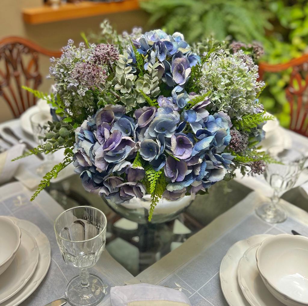 Centro de Mesa con Hydrangeas en Tonos Azules y Follajes Artificiales en Base de Vidrio Tipo Espejo.