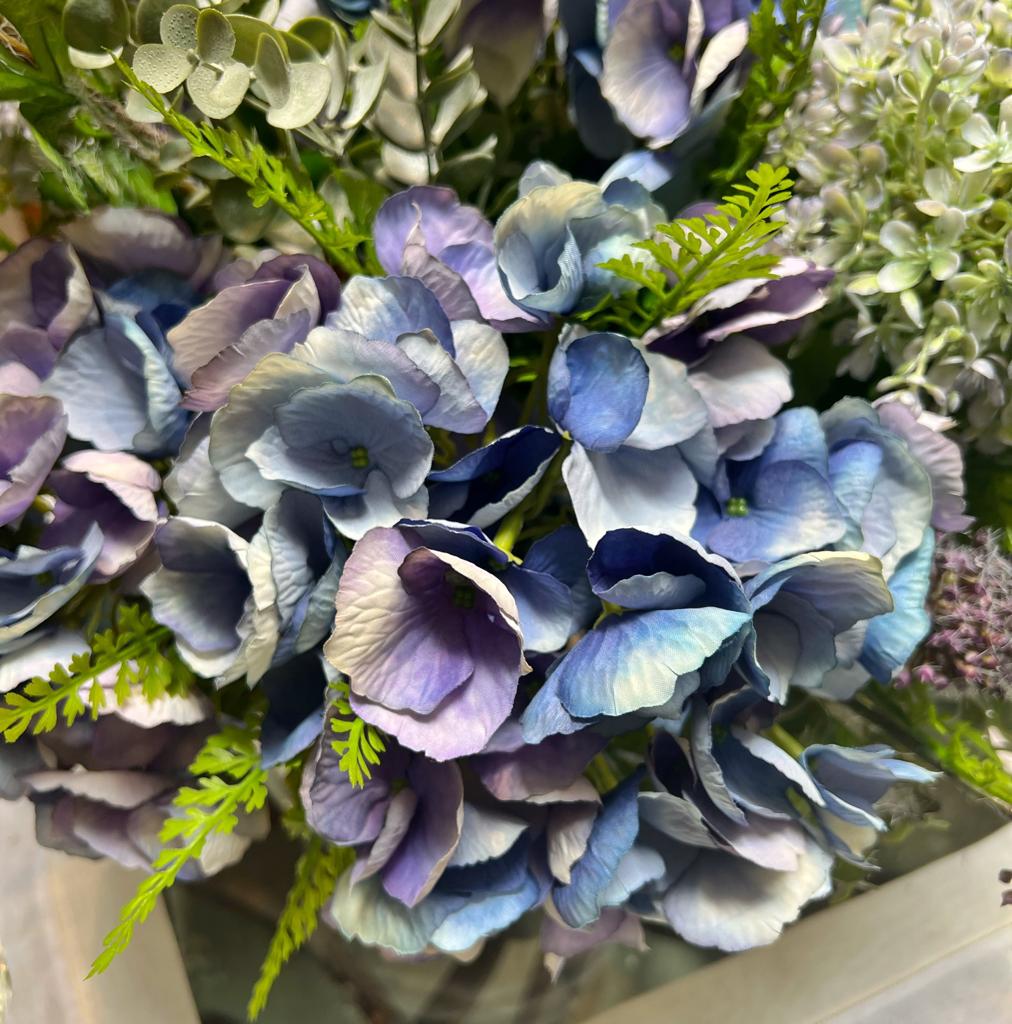 Centro de Mesa con Hydrangeas en Tonos Azules y Follajes Artificiales en Base de Vidrio Tipo Espejo.