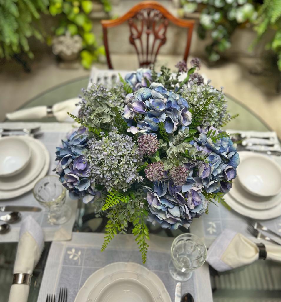 Centro de Mesa con Hydrangeas en Tonos Azules y Follajes Artificiales en Base de Vidrio Tipo Espejo.