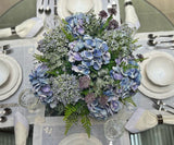 Centro de Mesa con Hydrangeas en Tonos Azules y Follajes Artificiales en Base de Vidrio Tipo Espejo.