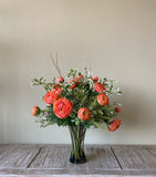 Arreglo Floral con Ranunculus Naranjas y Follajes Artificiales en Florero de Vidrio