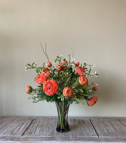 Arreglo Floral con Ranunculus Naranjas y Follajes Artificiales en Florero de Vidrio