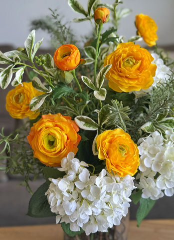 Arreglo Floral con Ranunculus Amarillos y Follajes Artificiales en Florero de Vidrio