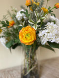 Arreglo Floral con Ranunculus Amarillos y Follajes Artificiales en Florero de Vidrio