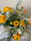 Arreglo Floral con Ranunculus Amarillos y Follajes Artificiales en Florero de Vidrio