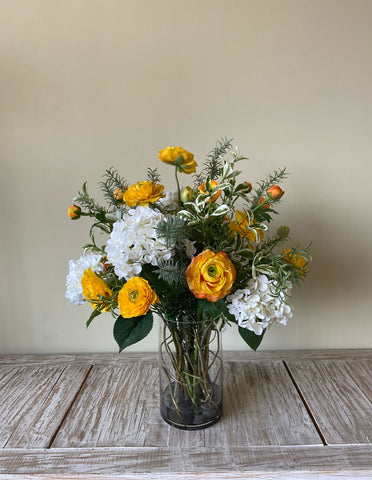 Arreglo Floral con Ranunculus Amarillos y Follajes Artificiales en Florero de Vidrio