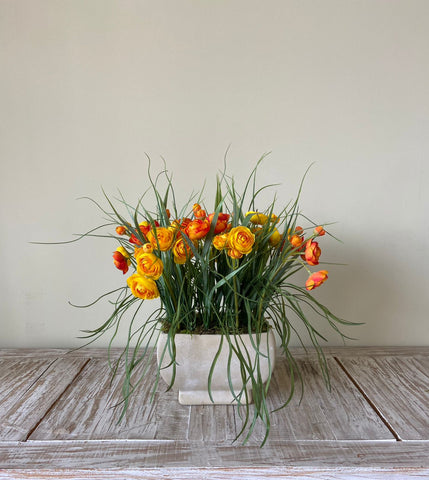 Arreglo Floral con Ranunculus Naranjas y Amarillos con Follajes Artificiales en Base Rectangular