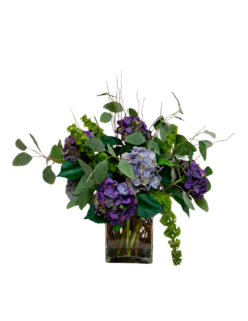 Arreglo Floral 3/4 con Hidrangeas Lilas y Follajes Artificiales en Base Cuadrada de Vidrio con Agua Acrílica