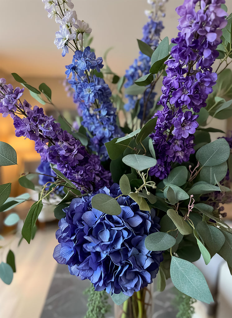 Arreglo Floral de Delphiniums e Hidrangeas con Follajes Artificiales en Cilindro de Vidrio